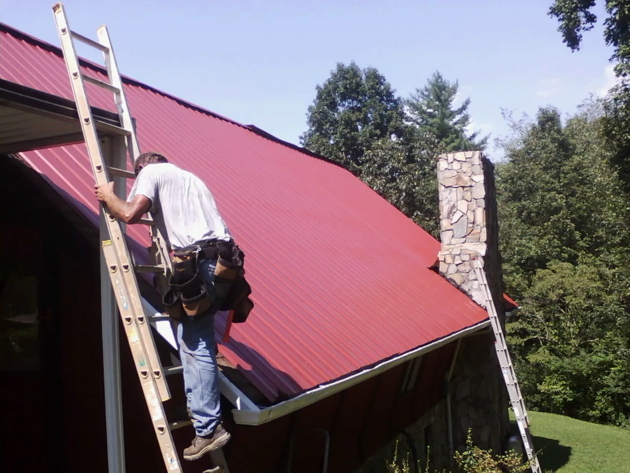Roofer on a ladder beside a red metal roof and stone chimney — Soffit & Fascia Repair by Roofing Brothers in Mukwonago